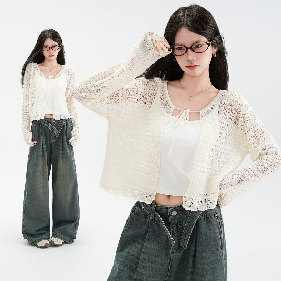 A woman wearing a cream open-knit cardigan patterned with fine pointelle rows and tied at the neck, worn over a white camisole with dark green wide leg jeans, her hair caught in the wind.