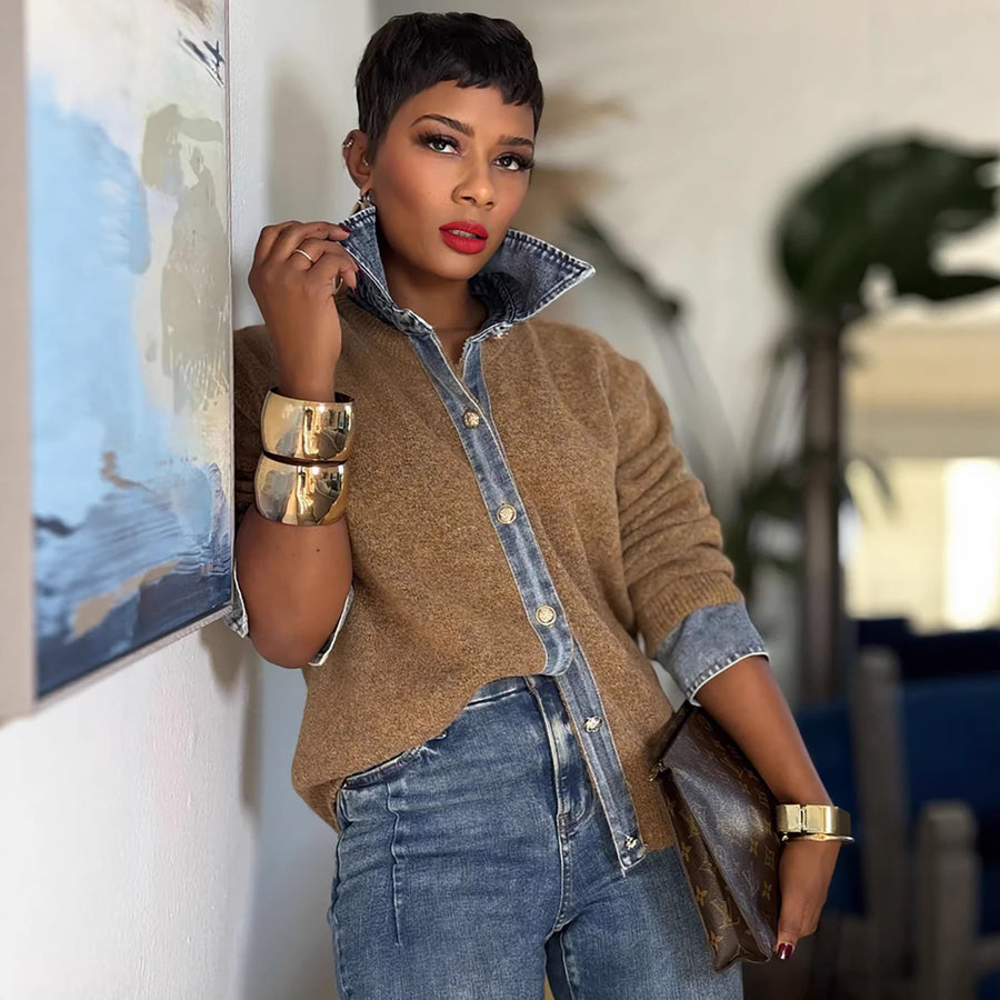 A woman wearing a grey knitted jumper finished with a denim shirt collar, a denim button placket and denim cuffs, giving the look of a shirt layered under a sweater, standing beside a framed picture.