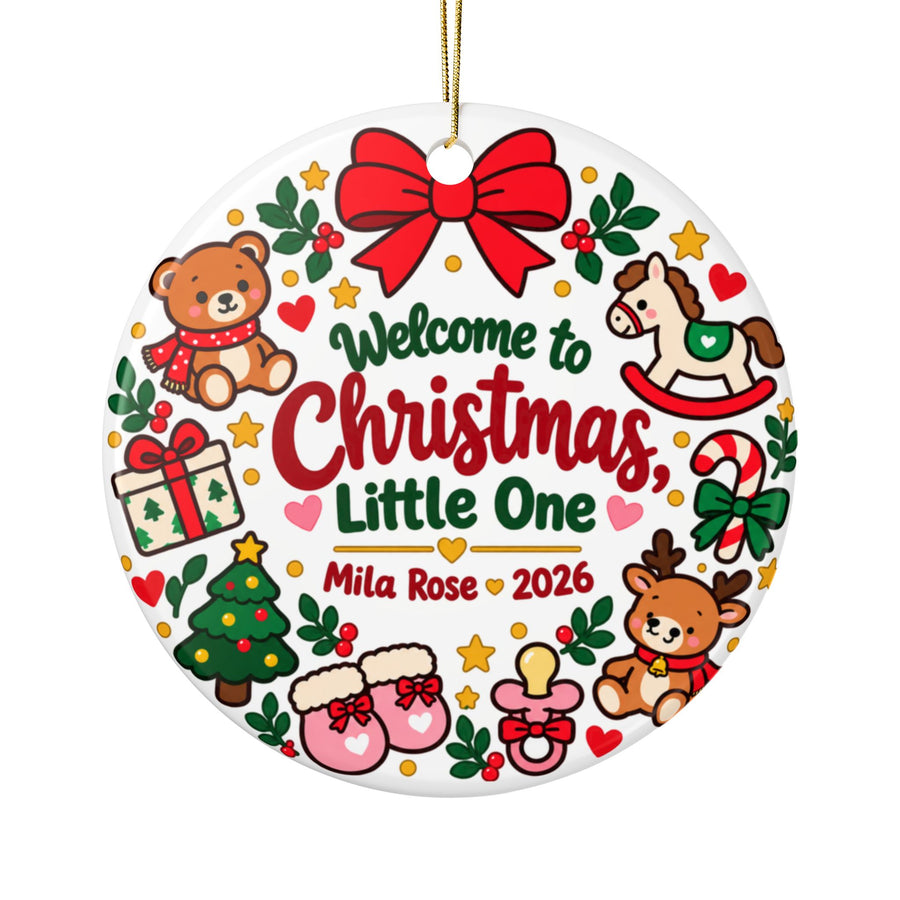 A round white ceramic Christmas ornament on a gold string, printed with "Welcome to Christmas, Little One" in green and red lettering above a sample baby name and "2026", ringed by a red bow, a teddy bear, a rocking horse, a reindeer, a candy cane, baby booties, a pacifier, a present and a tree.