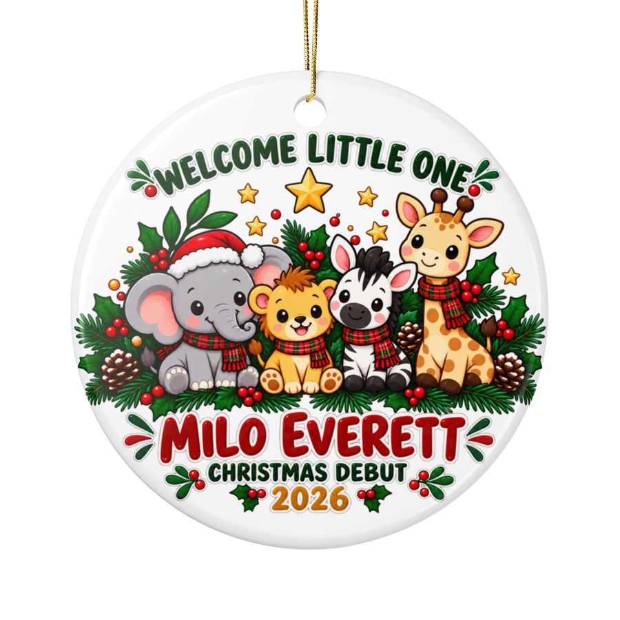 A round white ceramic Christmas ornament on a gold string, printed with "Welcome Little One" in green lettering over a baby elephant in a Santa hat, a lion cub, a zebra and a giraffe in plaid scarves among evergreen and pinecones, above a sample baby name in red, "Christmas Debut" and "2026".