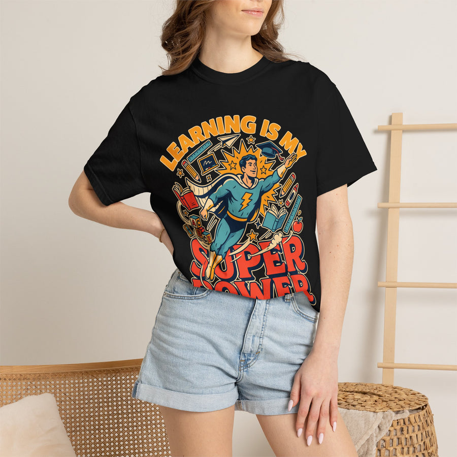 A black T-shirt printed with a comic-style teacher superhero flying among books and school supplies under Learning Is My Super Power.