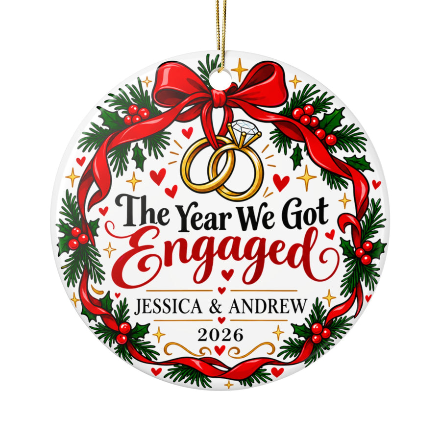 A round white ceramic Christmas ornament on a gold string, printed with two linked gold rings, one with a diamond, hanging from a red bow on a holly wreath, around "The Year We Got Engaged" in black and red lettering, sample couple names and "2026".