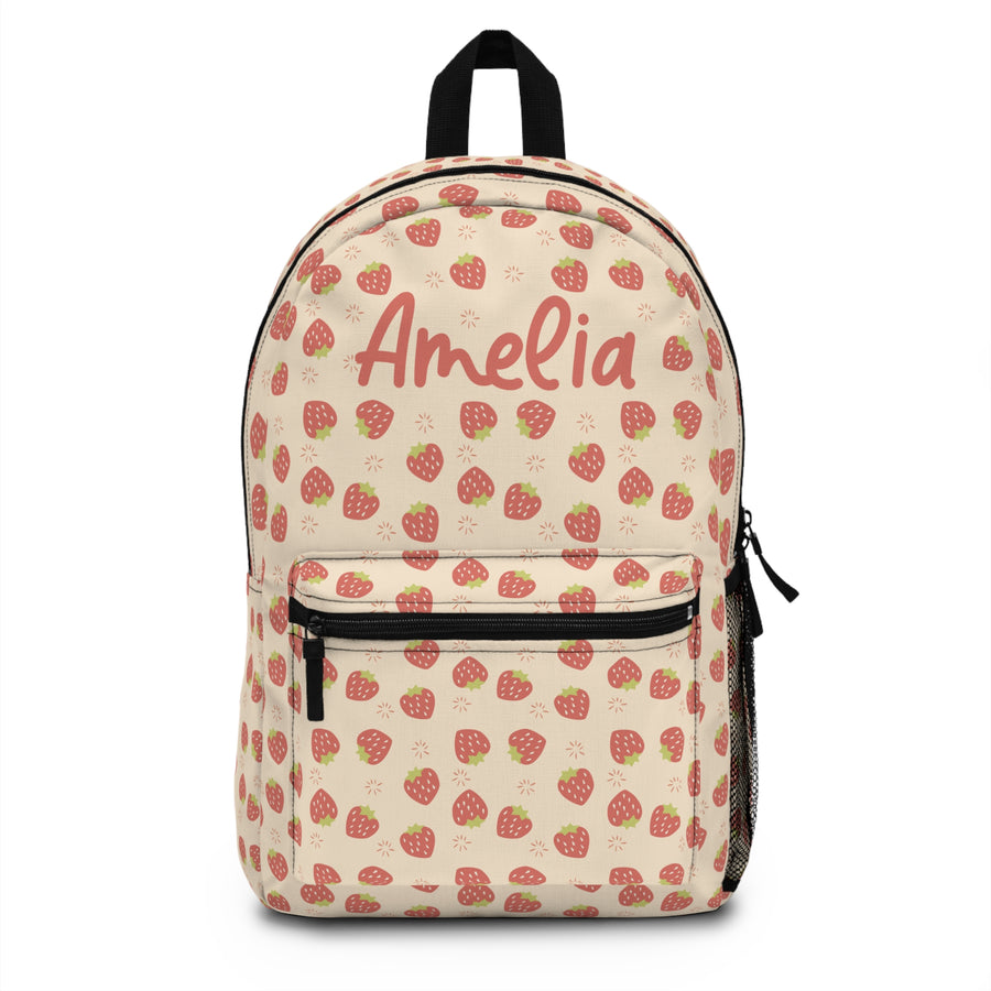A cream backpack covered in an all-over red strawberry print, black straps and zippers, and the sample name "Amelia" printed in coral script on the front, resting on a wooden desk.