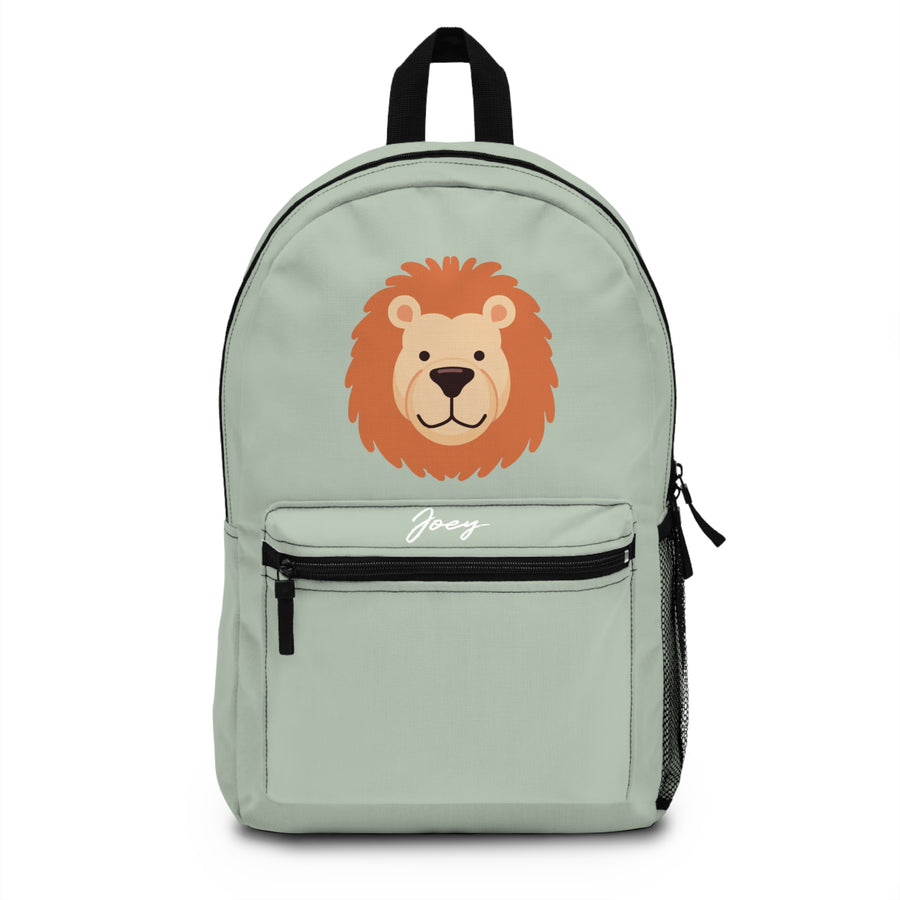A kids backpack with a black top handle, front zip pocket and mesh side pocket, sage green with a large friendly lion face with an orange mane and the sample name "Joey" in white script on the pocket.