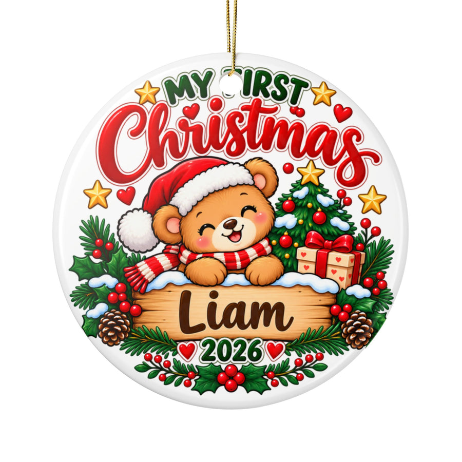 A round white ceramic Christmas ornament on a gold string, printed with "My First Christmas" in green and red lettering over a smiling teddy bear in a Santa hat and striped scarf, peeking over a wooden sign with a sample baby name and "2026", with a decorated tree, a present, pinecones and holly.
