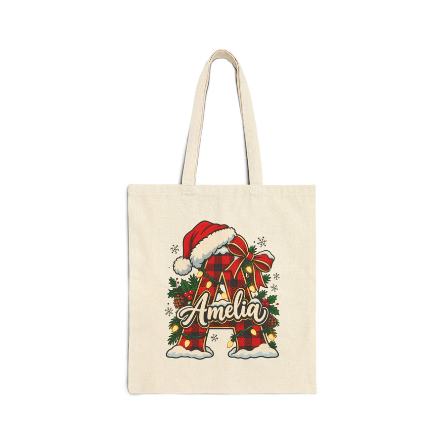 A natural cotton canvas tote bag with canvas handles, printed with a red and black buffalo plaid initial wearing a Santa hat with a red bow, holly, pinecones and string lights, and a sample name in white script across it, in front of a frosted Christmas tree and presents.