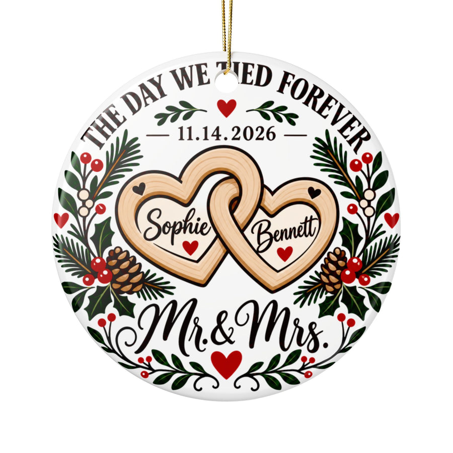 A round white ceramic Christmas ornament on a gold string, printed with "THE DAY WE TIED FOREVER" arched over a sample wedding date, two linked wooden-look hearts with sample names, holly, pinecones and red berries, and "Mr. & Mrs." in black script.