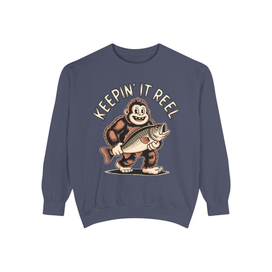 White sweatshirt printed with a grinning bigfoot carrying a large open-mouthed fish under one arm, headed Keepin It Reel in an arc of outlined capitals curving above his head.