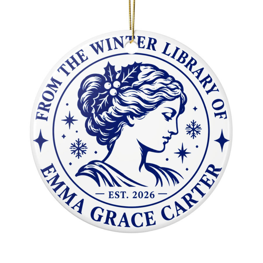 A round white ceramic Christmas ornament on a gold string, printed with "FROM THE WINTER LIBRARY OF" and a sample name in navy serif lettering around a classic portrait of a woman with holly in her hair, snowflakes, stars and "EST. 2026".