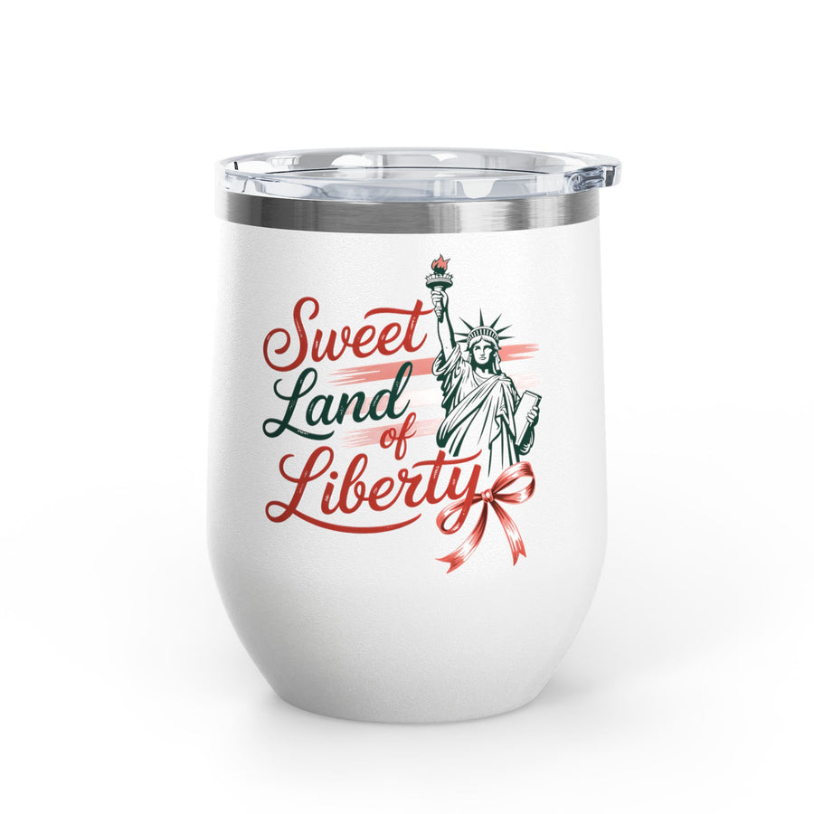 A white stainless steel wine tumbler with a clear push-on lid, printed with a red and green "Sweet Land of Liberty" design featuring the Statue of Liberty and a bow, on a wooden table.