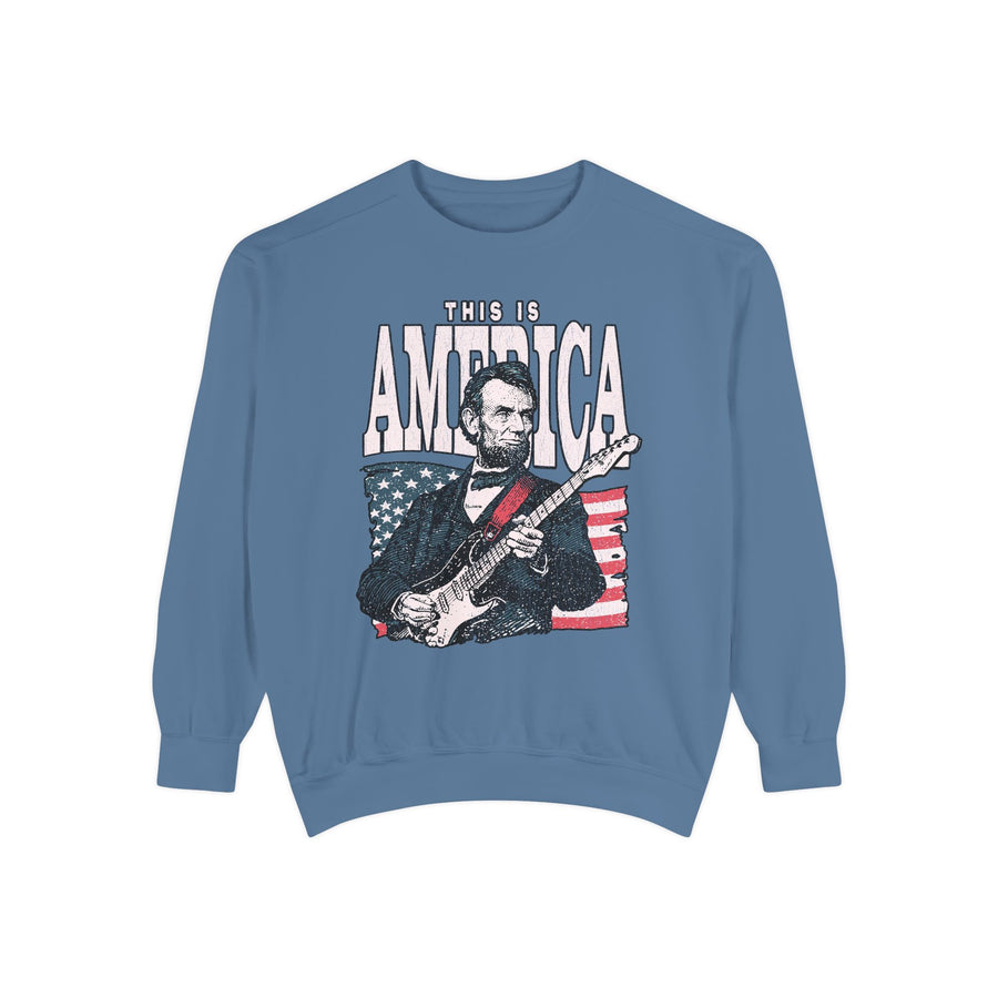 A blue crewneck sweatshirt printed with an illustration of Abraham Lincoln playing an electric guitar in front of an American flag under This Is America.