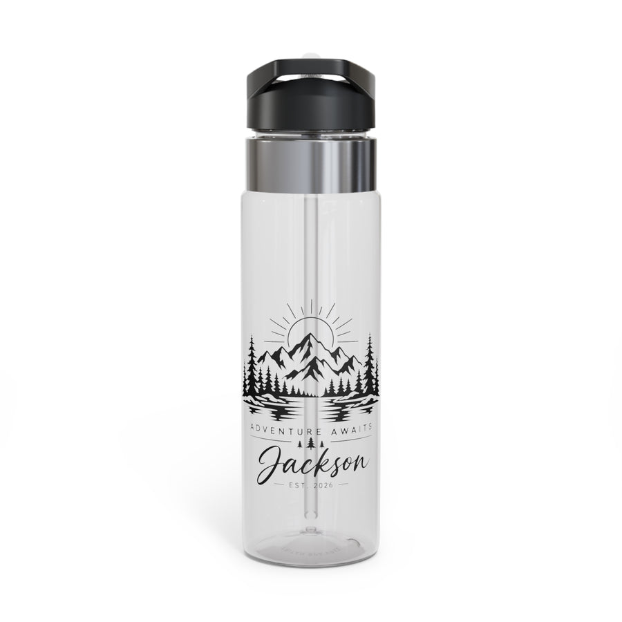 A clear water bottle with a black flip-top lid, stainless band and straw, printed with black mountains, pine trees, a lake and a rising sun above Adventure Awaits and the sample name "Jackson".