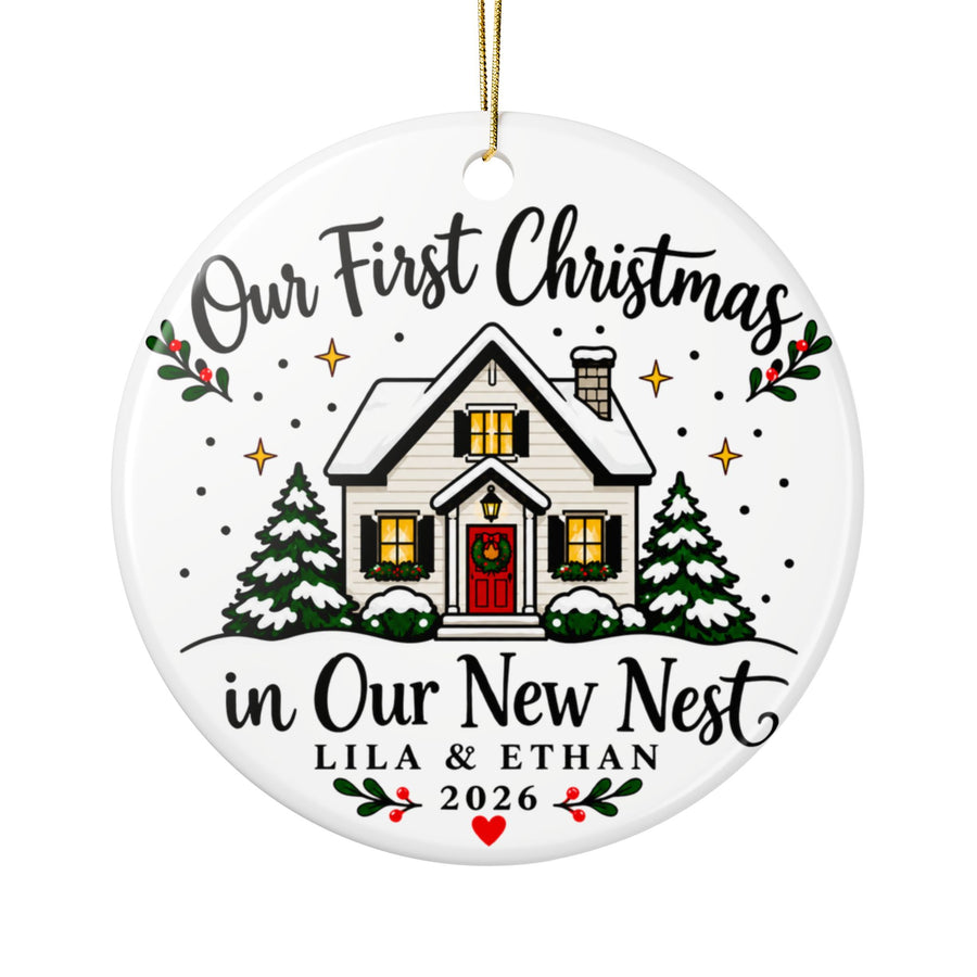 A round white ceramic Christmas ornament on a gold string, printed with "Our First Christmas in Our New Nest" in black script around a snow-covered white house with a red door, glowing windows and snowy pine trees, with sample couple names, "2026" and a red heart.