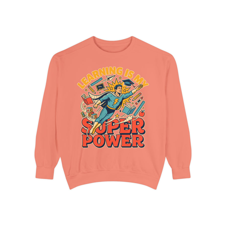 White sweatshirt printed with a comic-style teacher flying like a superhero in a cape and mortarboard through books and pencils, headed Learning Is My Super Power in orange.