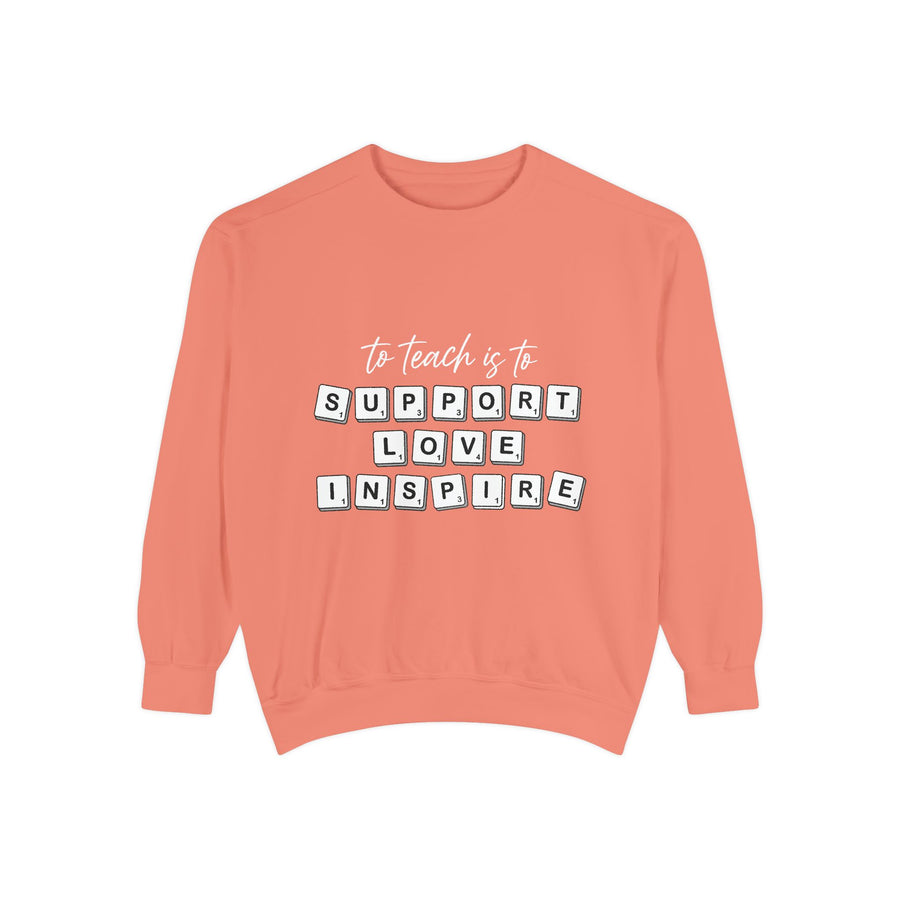 Grey sweatshirt printed with the words to teach is to in slim script above Support Love Inspire, each word spelled out in individual Scrabble-style letter tiles laid out as if on a board.