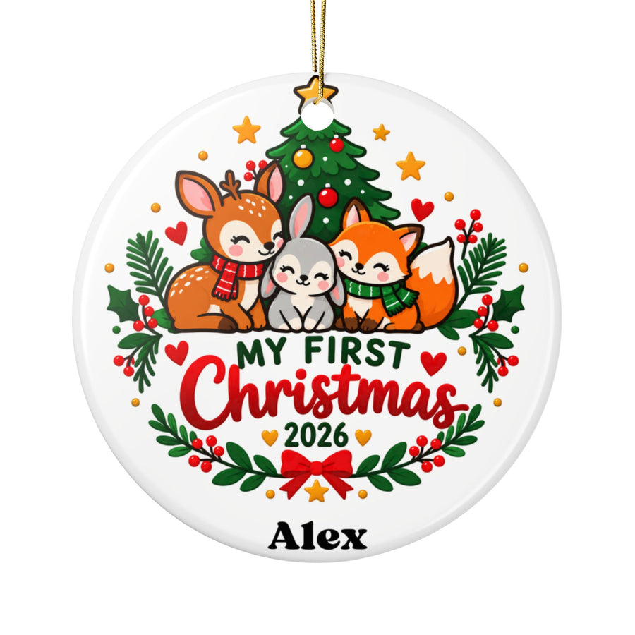 A round white ceramic Christmas ornament on a gold string, printed with a deer, a gray bunny and a fox in knit scarves cuddled under a decorated Christmas tree, above "My First Christmas" in green and red lettering, "2026", a holly garland with a red bow and a sample baby name.