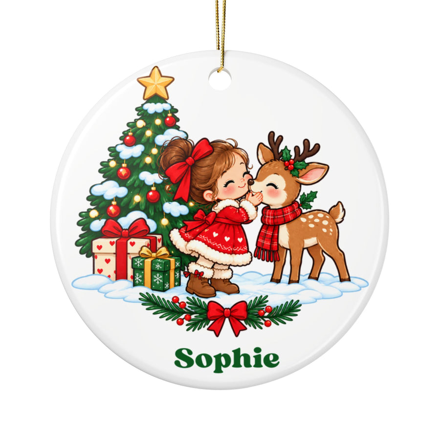 A round white ceramic Christmas ornament on a gold string, printed with a little girl in a red dress and hair bow kissing a deer in a plaid scarf beside a decorated Christmas tree and presents in the snow, above a holly garland and a sample girl's name in green.