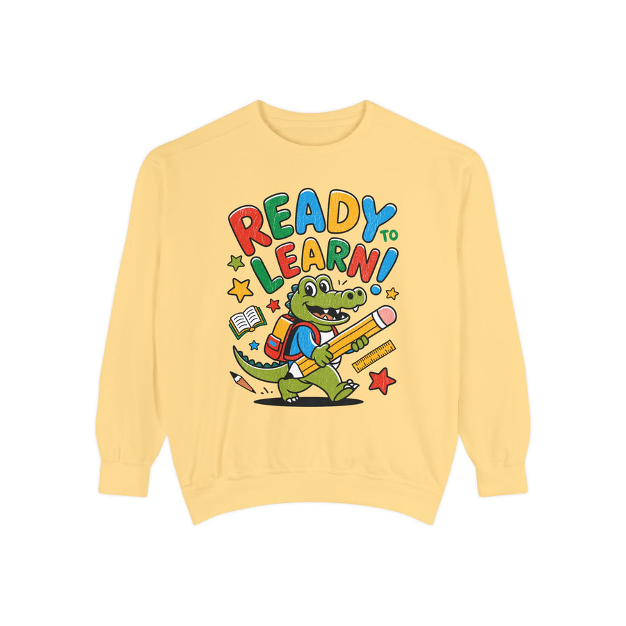 A green crewneck sweatshirt printed with a cartoon alligator in a backpack carrying a pencil under Ready to Learn! in rainbow letters.