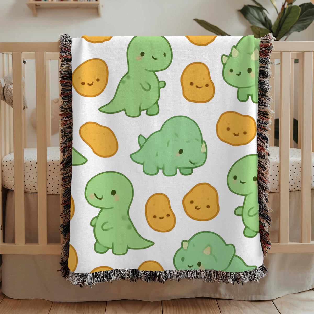 White fringed woven blanket draped over a wooden crib, printed with smiling pale green cartoon dinosaurs and rounded golden nugget shapes, each drawn with a simple happy face.