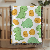 White fringed woven blanket draped over a wooden crib, printed with smiling pale green cartoon dinosaurs and rounded golden nugget shapes, each drawn with a simple happy face.
