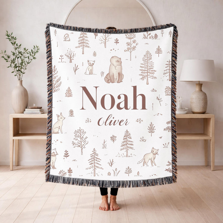White fringed woven blanket over a cream crib, printed with line drawn pine trees, a bear and foxes in soft brown, with the name Noah in large serif and Oliver in script beneath.
