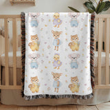 Fringed woven baby blanket draped over a wooden crib, printed with rows of watercolour koalas in blue overalls, ducklings in purple dresses, fawns and squirrels holding tiny posies.