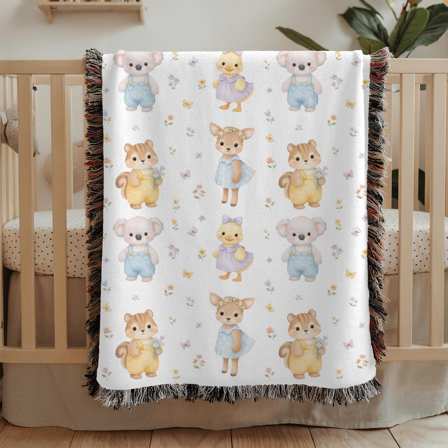 Fringed woven baby blanket draped over a wooden crib, printed with rows of watercolour koalas in blue overalls, ducklings in purple dresses, fawns and squirrels holding tiny posies.