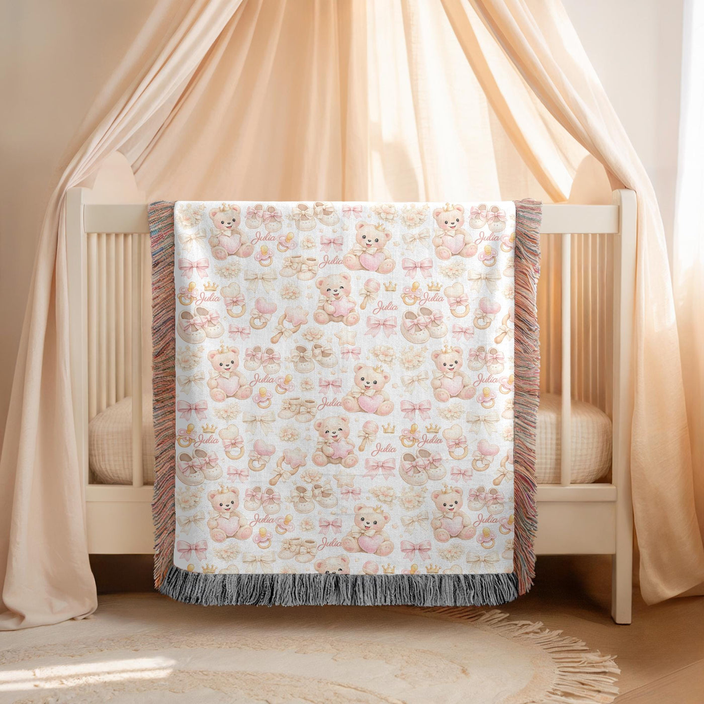 White fringed woven blanket over a cream crib, printed with pink crowned teddy bears holding hearts among bows, booties, rattles and pacifiers, with the name Julia in pink script.