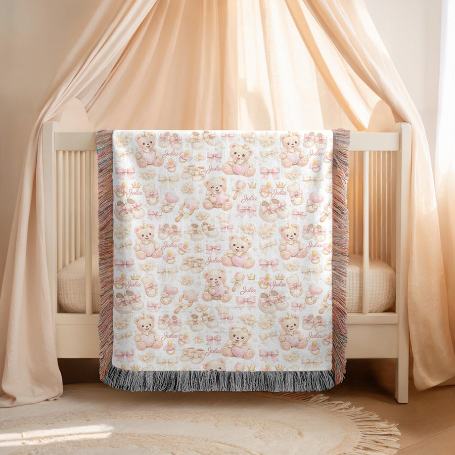 White fringed woven blanket over a cream crib, printed with pink crowned teddy bears holding hearts among bows, booties, rattles and pacifiers, with the name Julia in pink script.