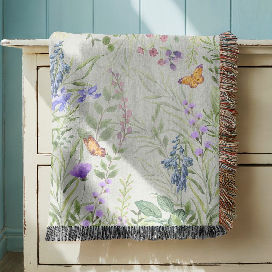 Pale green fringed woven blanket printed with watercolour wildflowers, bluebells, purple irises and slender pink stems, with two orange butterflies resting among the leaves.