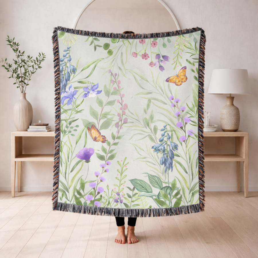 Pale green fringed woven blanket printed with watercolour wildflowers, bluebells, purple irises and slender pink stems, with two orange butterflies resting among the leaves.