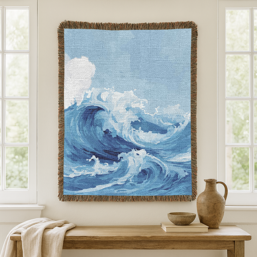 Blue fringed woven blanket painted with a large ocean wave curling over and breaking into white foam, its crest rising against a pale grey sky above the open water beyond.