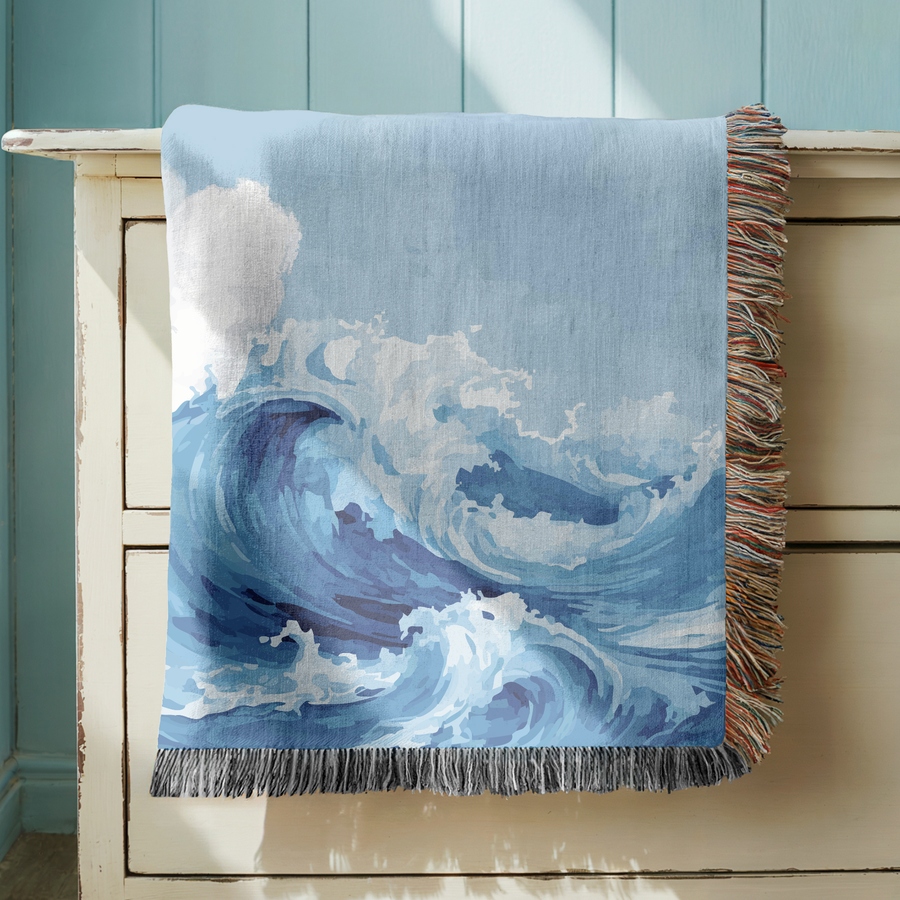 Blue fringed woven blanket painted with a large ocean wave curling over and breaking into white foam, its crest rising against a pale grey sky above the open water beyond.