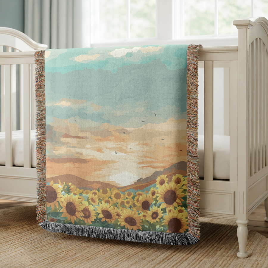 Fringed woven blanket printed with a field of open sunflowers below rolling hills, a wide sunset sky of teal and apricot cloud above them and small birds crossing the light.