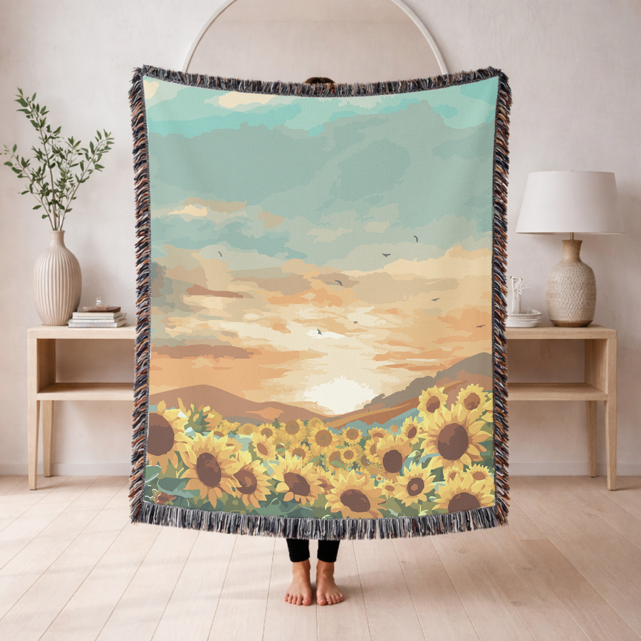 Fringed woven blanket printed with a field of open sunflowers below rolling hills, a wide sunset sky of teal and apricot cloud above them and small birds crossing the light.