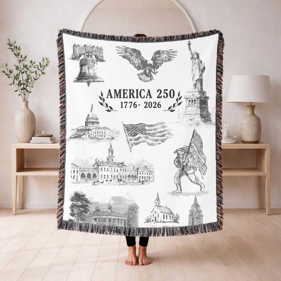 Cream fringed woven blanket headed America 250, 1776 to 2026, with engraved views of the Liberty Bell, the Statue of Liberty, the Capitol, a waving flag and a marching soldier.