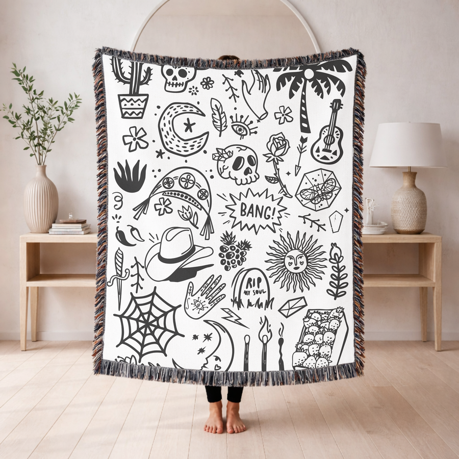 White fringed woven blanket covered in small black doodles: skulls, a cactus, a crescent moon, a palm tree, a guitar, a rose, a spiderweb, a cowboy hat and a sun with a face.