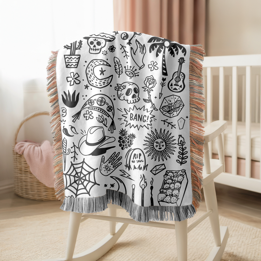 White fringed woven blanket covered in small black doodles: skulls, a cactus, a crescent moon, a palm tree, a guitar, a rose, a spiderweb, a cowboy hat and a sun with a face.