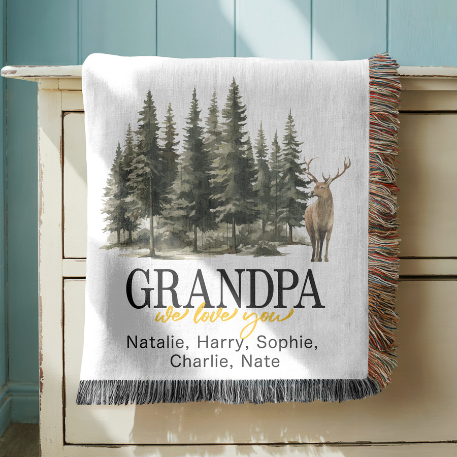 White fringed woven blanket printed with a watercolour pine forest and a standing antlered stag, reading Grandpa above we love you in gold and five names in a line beneath.