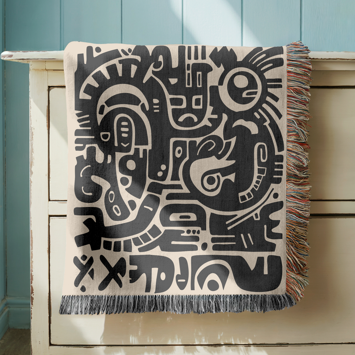 Cream fringed woven blanket printed with bold black abstract shapes packed edge to edge, among them stylised faces, eyes and rounded geometric forms in a rough hand-cut style.