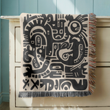 Cream fringed woven blanket printed with bold black abstract shapes packed edge to edge, among them stylised faces, eyes and rounded geometric forms in a rough hand-cut style.