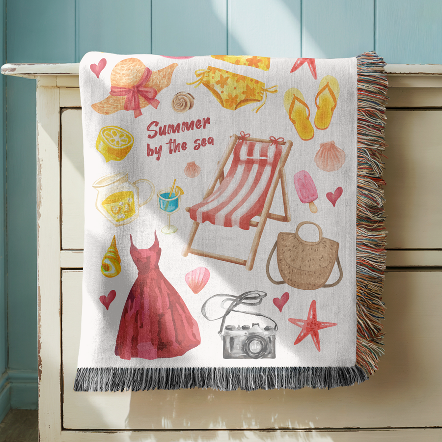 White fringed woven blanket reading Summer by the Sea among watercolour holiday things: a sun hat, a striped deck chair, flip-flops, an ice lolly, a straw bag and a camera.