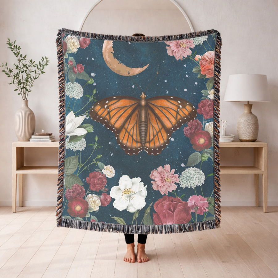 Deep blue fringed woven blanket printed with a large orange butterfly at its centre beneath a crescent moon, ringed with roses, peonies, magnolia and white hydrangea heads.