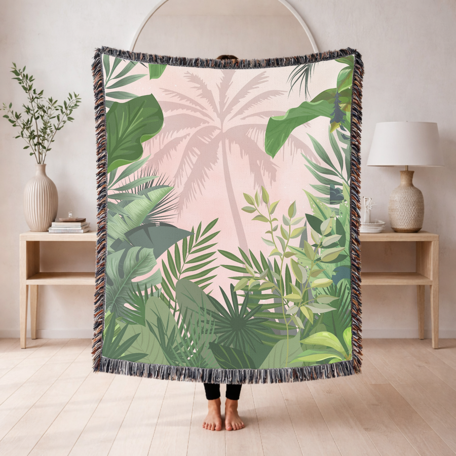 Fringed woven blanket in blush pink carrying the pale silhouette of a tall palm tree, framed on every side by layered green tropical leaves, monstera, fan palms and ferns.