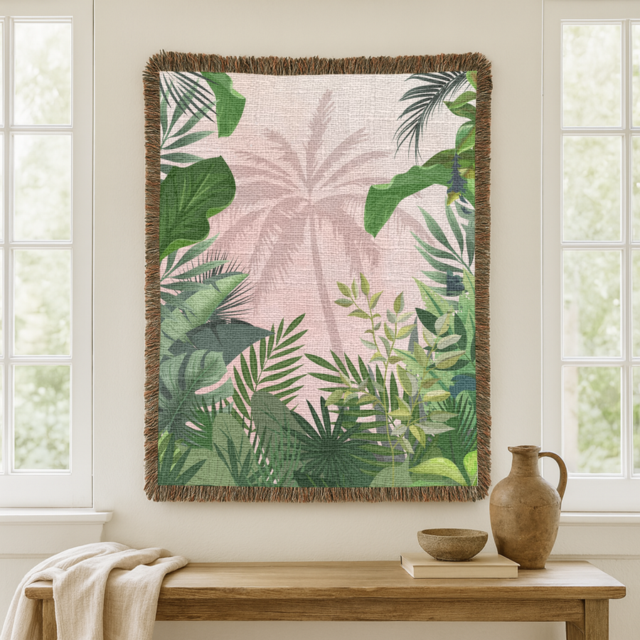 Fringed woven blanket in blush pink carrying the pale silhouette of a tall palm tree, framed on every side by layered green tropical leaves, monstera, fan palms and ferns.