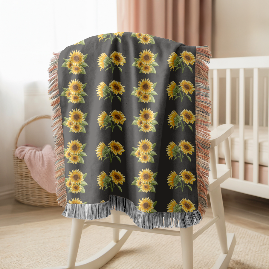 Charcoal fringed woven blanket printed with rows of watercolour sunflowers, their yellow petals and dark centres set against broad green leaves and stems on the dark ground.