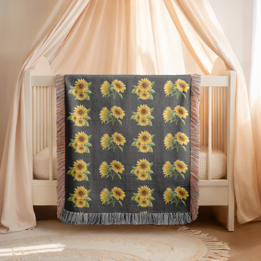 Charcoal fringed woven blanket printed with rows of watercolour sunflowers, their yellow petals and dark centres set against broad green leaves and stems on the dark ground.
