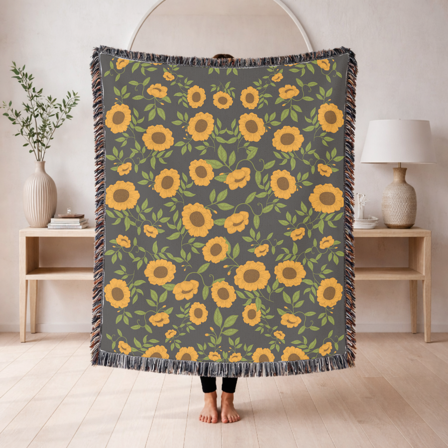 Grey fringed woven blanket covered in an even pattern of ochre sunflowers with bright green leaves and curling vines running between them across the whole face of the cloth.