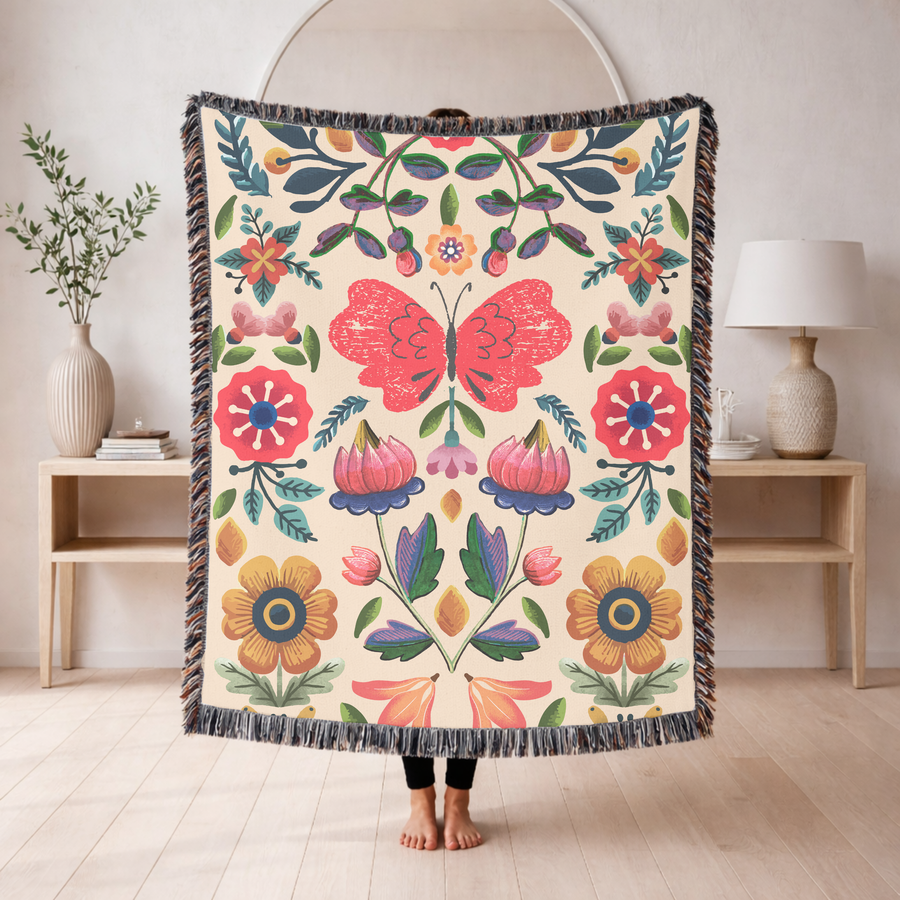 Cream fringed woven blanket printed with a symmetrical folk-art design centred on a large pink butterfly, with red and yellow flowers, blue leaves and tulips mirrored around it.