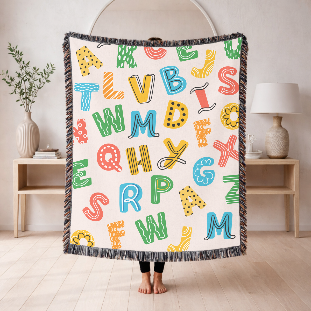 Cream fringed woven blanket scattered with big playful alphabet letters in green, blue, yellow and coral, each filled with dots, stripes or squiggles, with a few flowers between.